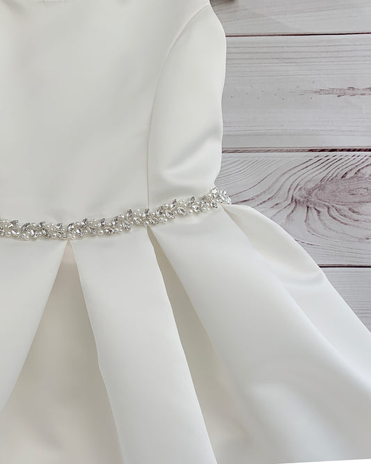White satin flower girl dress with a decorative belt on a wooden surface.