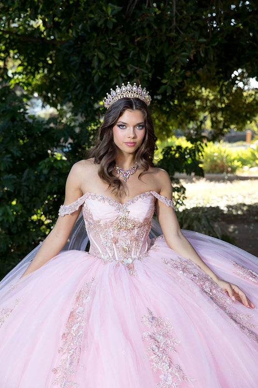 A woman wearing a blush pink off-shoulder ballgown with beading, rhinestones, and 3D flowers, featuring a golden embroidery and a detachable glitter cape.