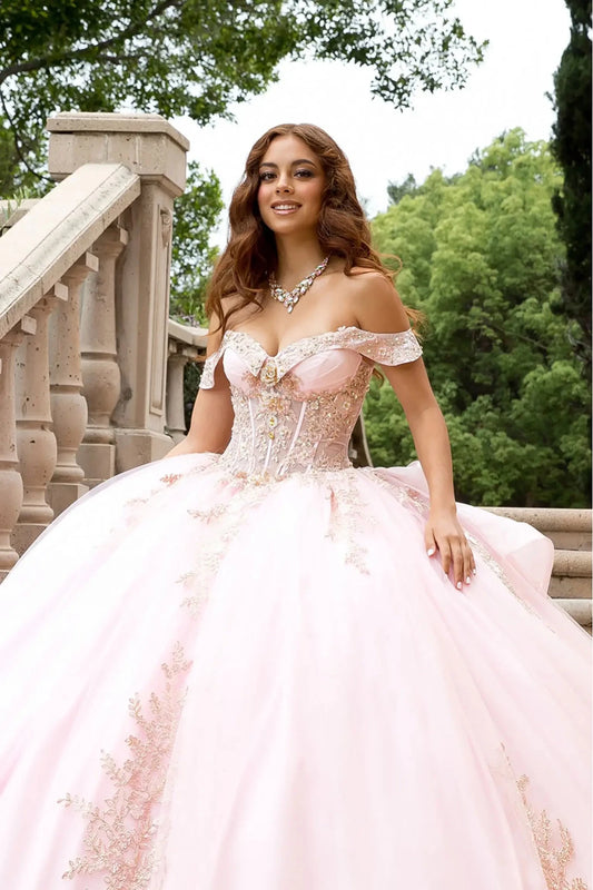 Model smiling while wearing the blush pink quinceañera gown, showcasing the sweetheart neckline and the jewelry-grade crystal embellishments on the corset. The sunlight catches the shimmery texture of the glitter-infused tulle fabric.
