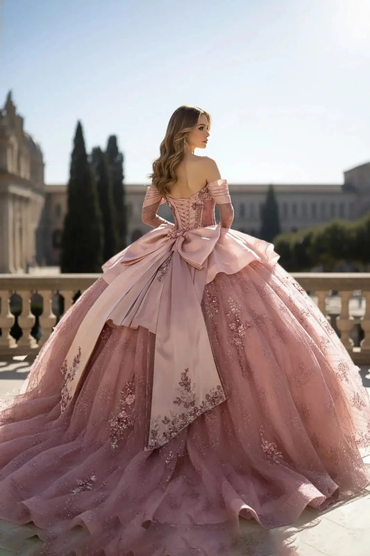 Back view of a blush pink off-the-shoulder ball gown featuring a corset-style back, a large decorative bow, and an expansive floral embroidered train, set on a stone terrace with tall cypress trees in the background.