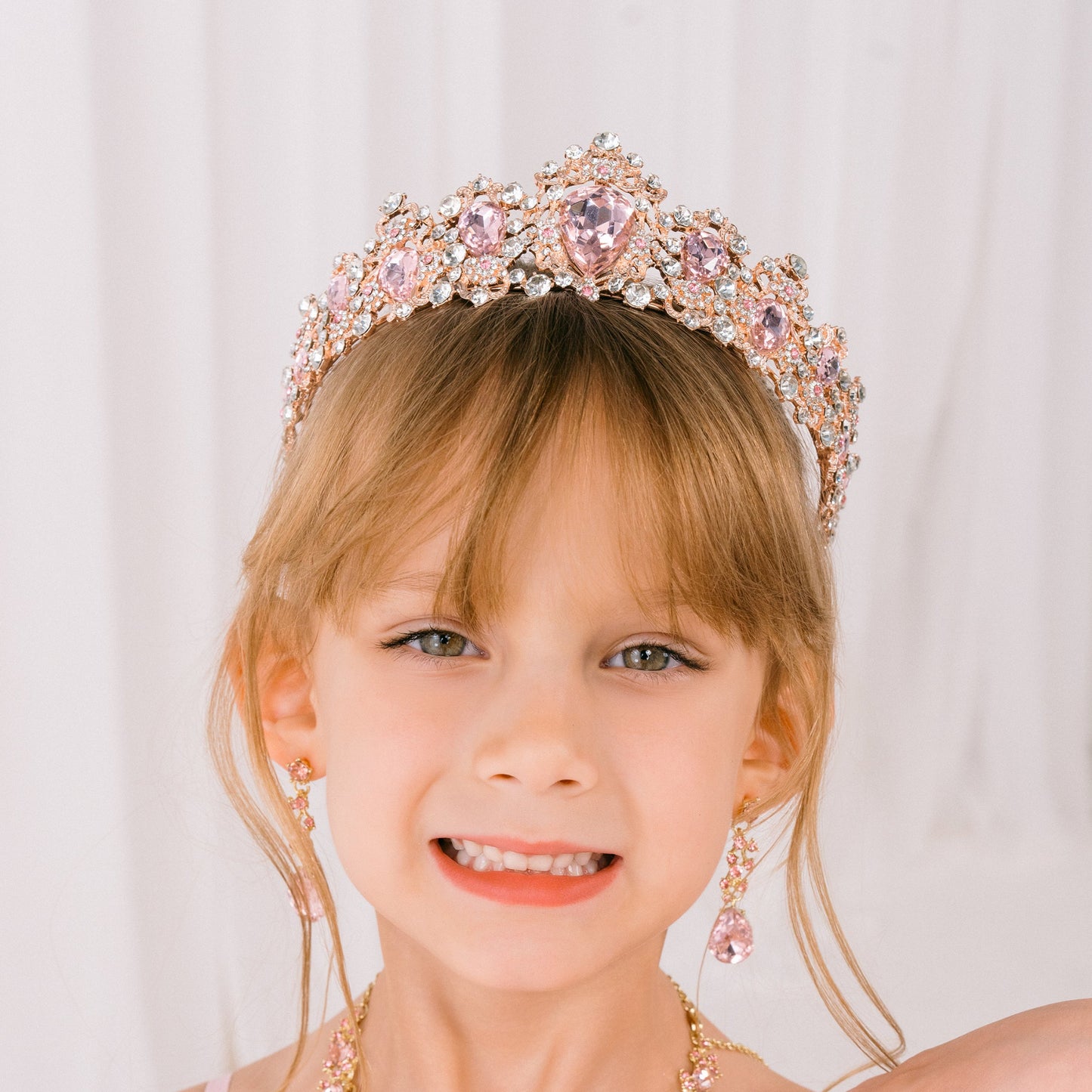 Young girl wearing a pink dress, tiara, and necklace against a white background