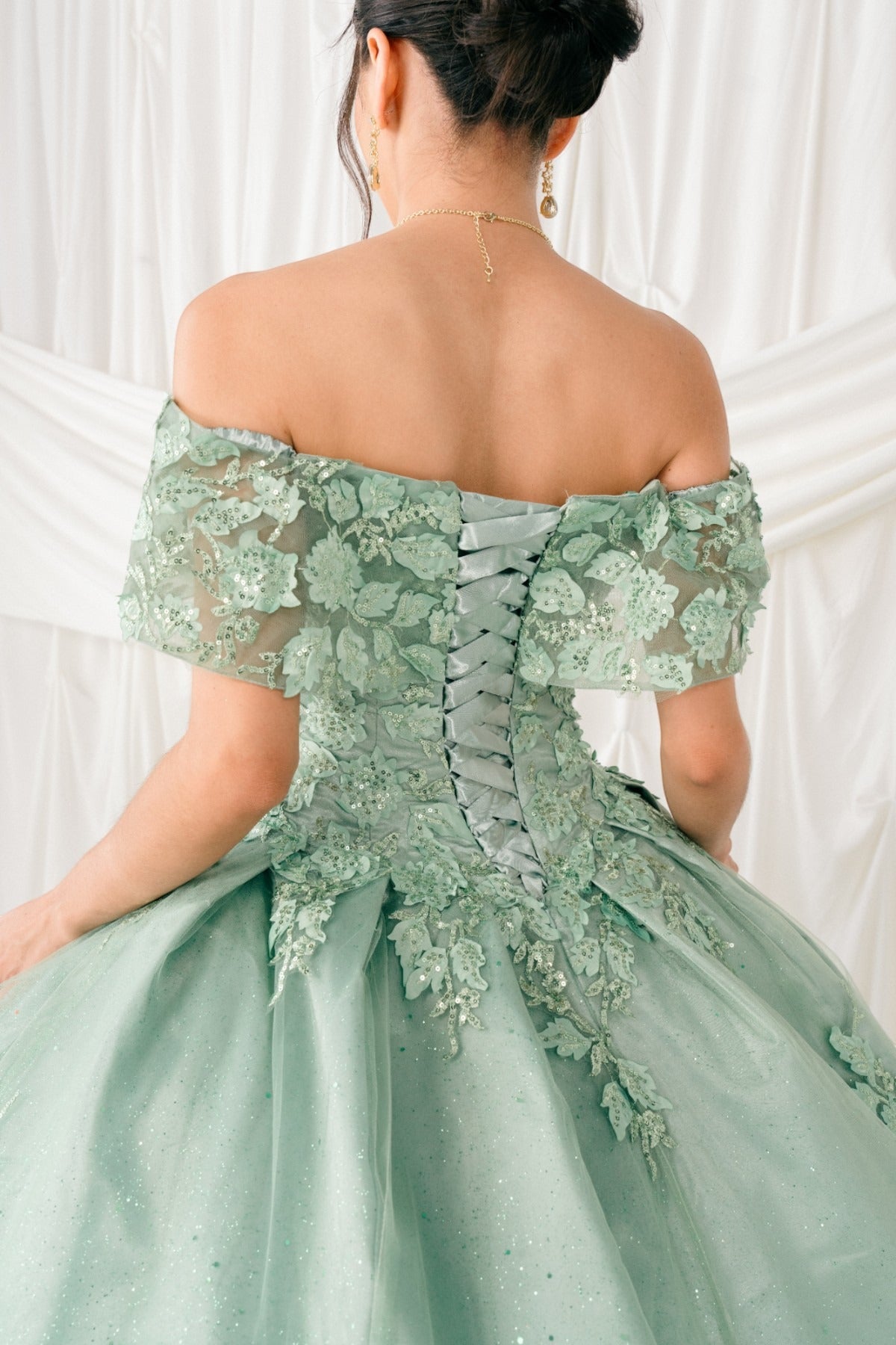 Bodice close-up highlights a Sage quinceanera dress with off-shoulder lace appliques, glitter accents, and voluminous skirt.