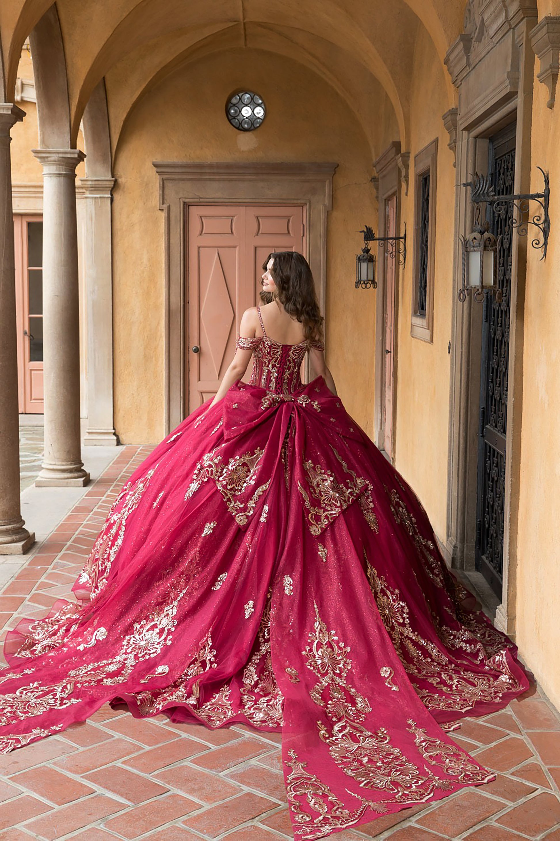 Woman in a red and gold embroidered gown standing in an outdoor setting with columns and a door.