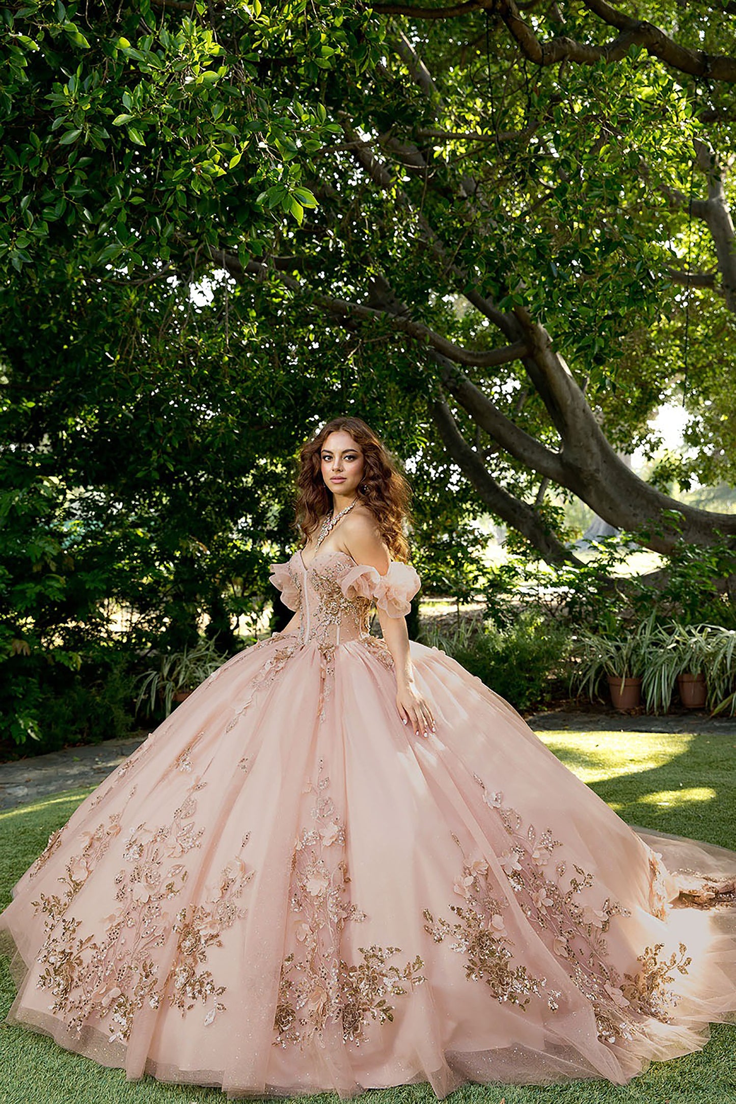 Woman in a pink and gold embroidered gown standing in a garden with trees and plants.