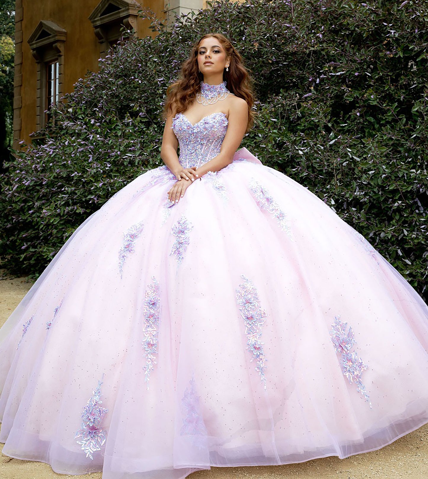 Woman in a pink and purple ball gown standing outdoors with greenery in the background