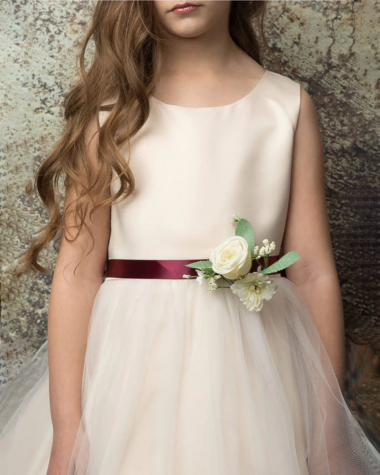 Young girl wearing a champagne dress with a floral burgundy sash against a textured wall.