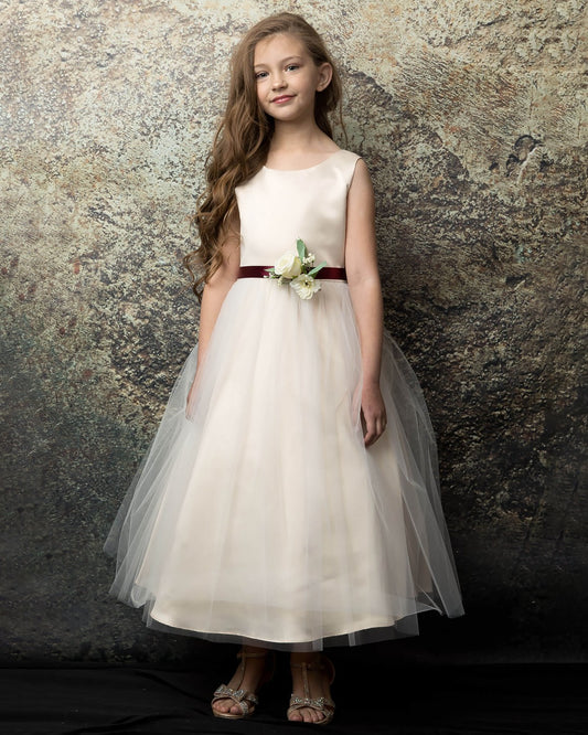 Young girl in a champagne dress with a red belt against a textured stone wall.