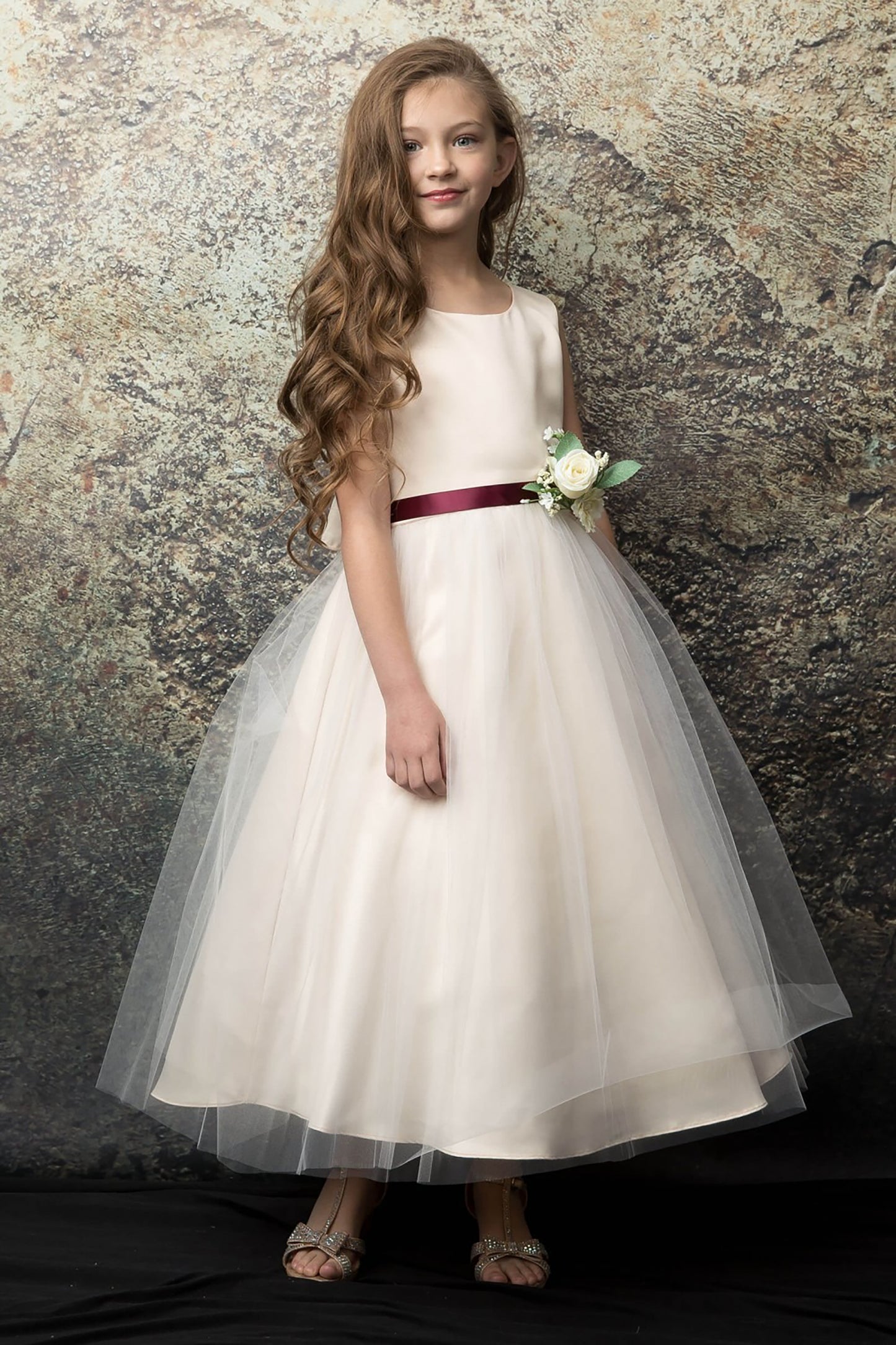 Young girl in a champagne dress with a burgundy belt against a textured stone wall.