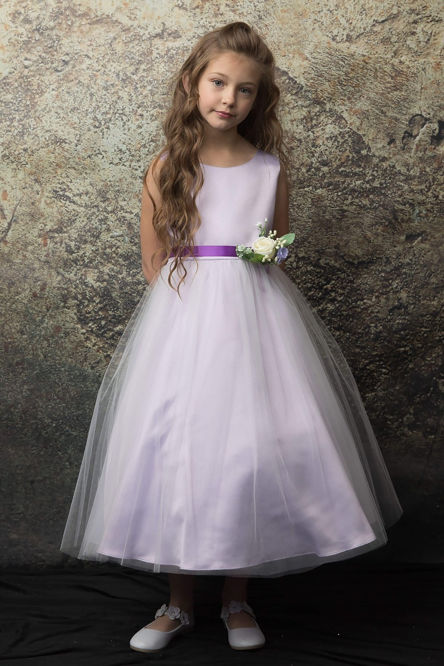 Young girl in a lilac dress with a purple belt and white flower against a textured wall.
