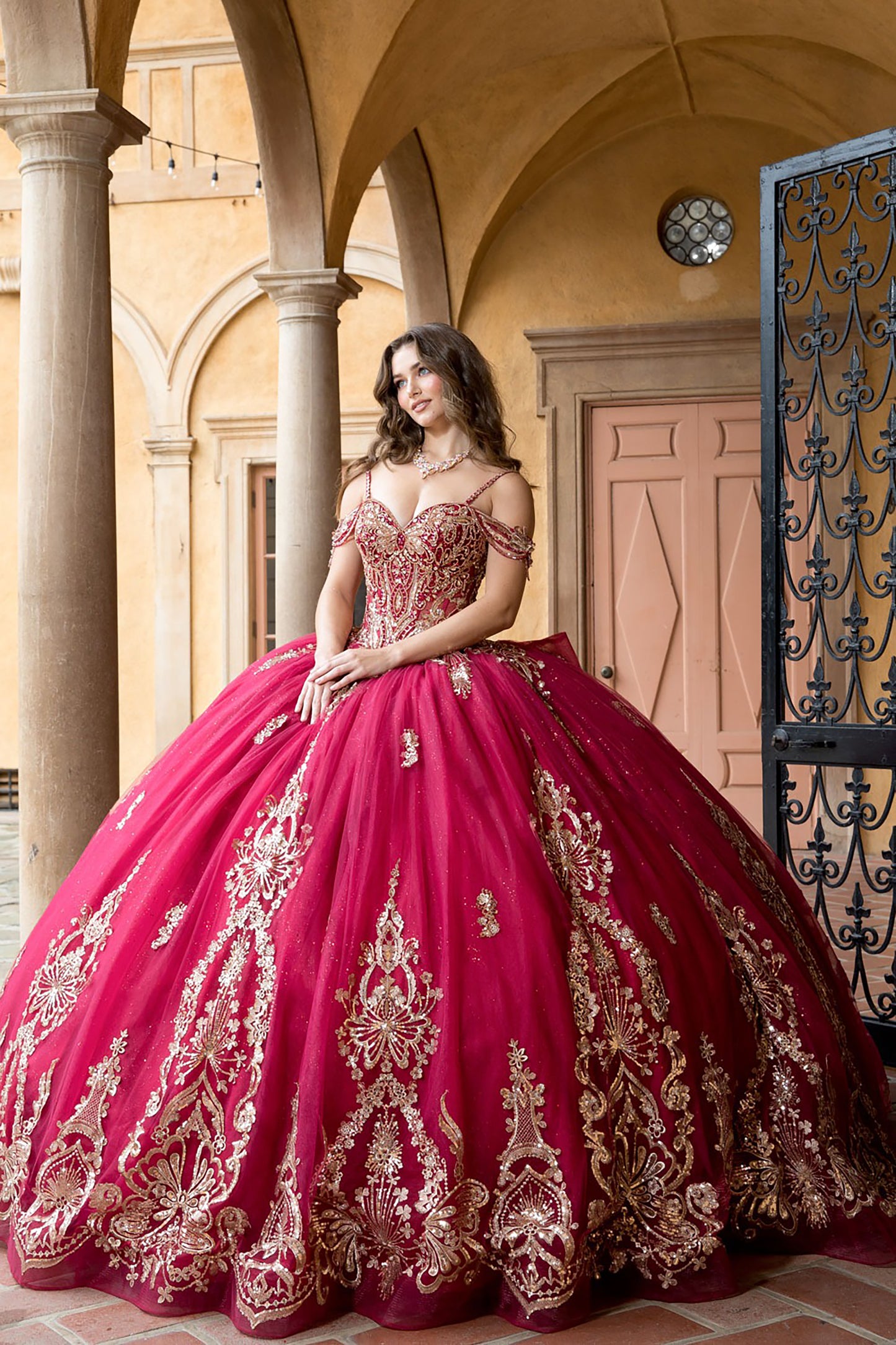 Woman in a red and gold embroidered ball gown standing in an outdoor setting with columns and a door.