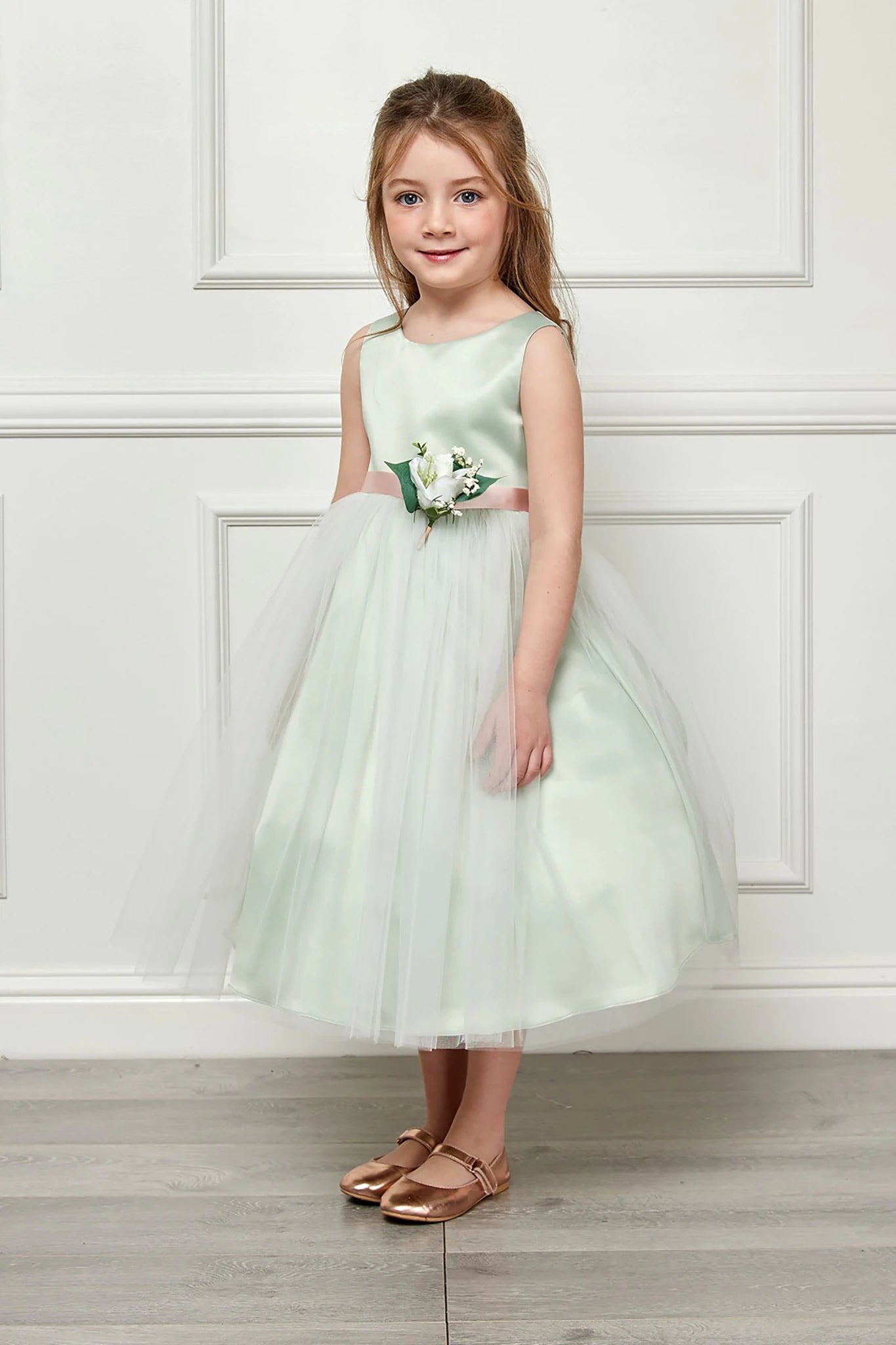 Young girl wearing a light green dress with a floral belt against a white paneled wall.