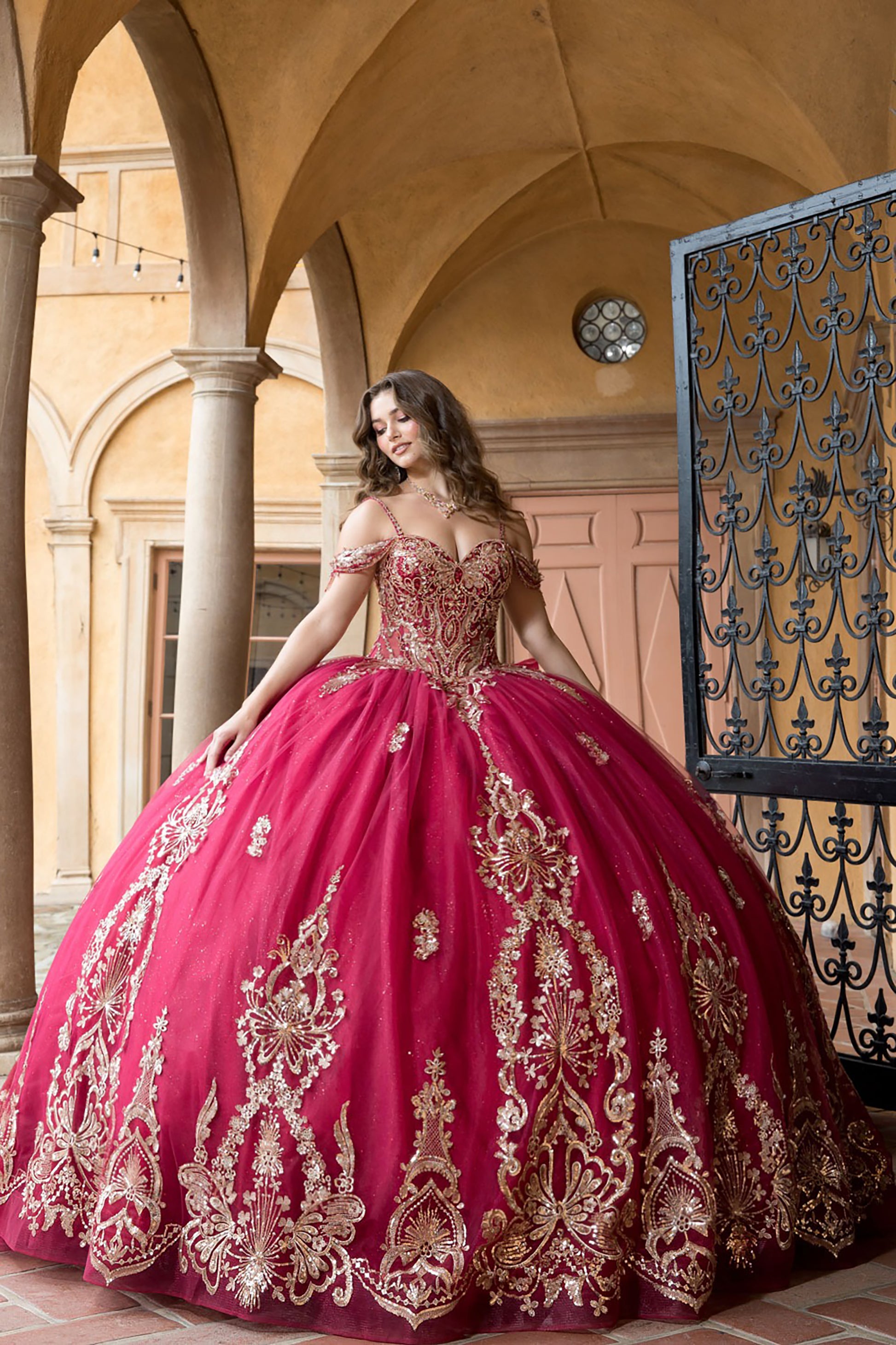 Woman in a red and gold embroidered ball gown standing in an elegant indoor setting.