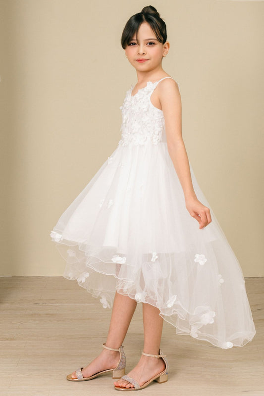 Young girl standing side-facing in white high-low tulle flower girl dress with 3D floral appliqué detailing and flowing layered high-low skirt