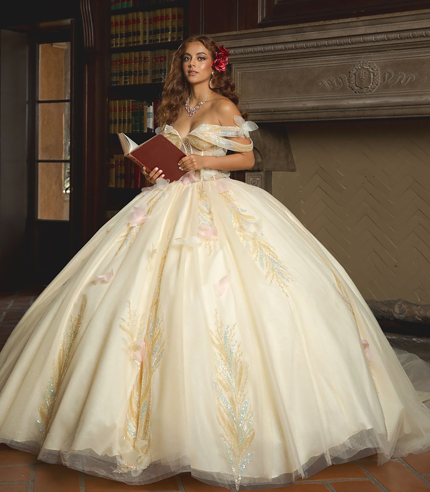 Woman in a white gown with floral details holding a red book in a room with a fireplace.