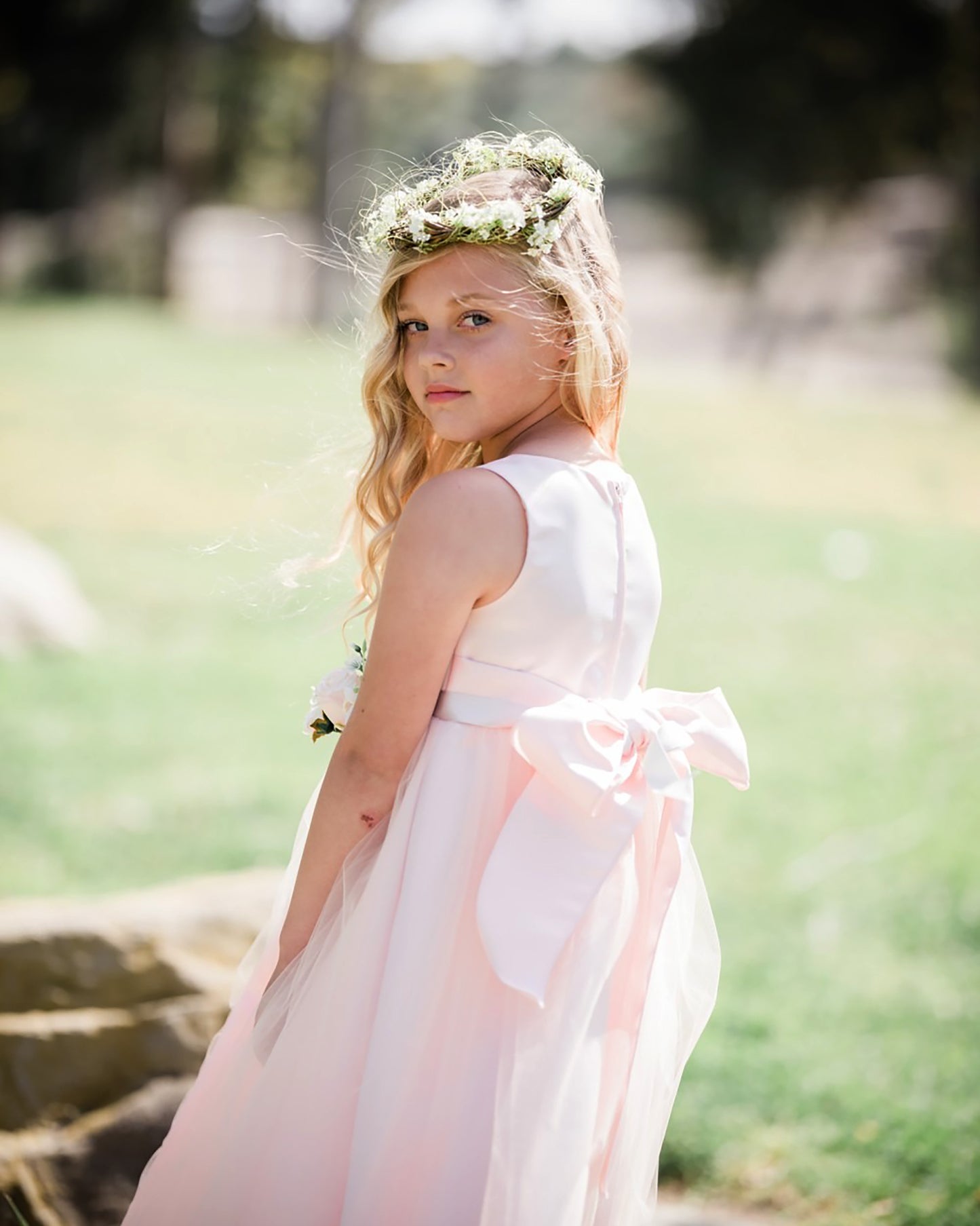 Young girl in a blush dress with a floral crown standing outdoors.