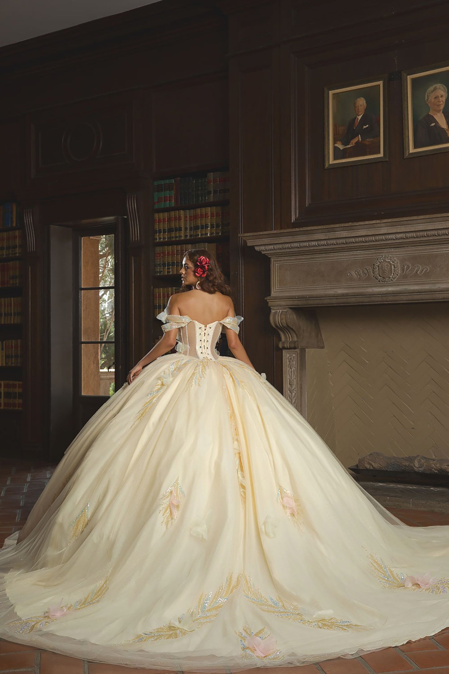 Woman in a white wedding dress with floral details standing in a room with wooden paneling and bookshelves.
