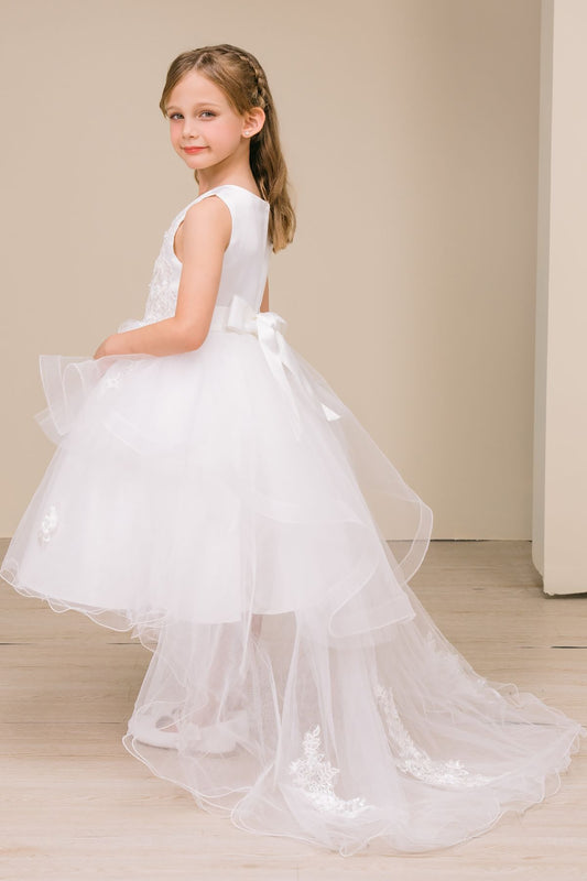 Young girl wearing a white dress with a bow, standing against a plain background
