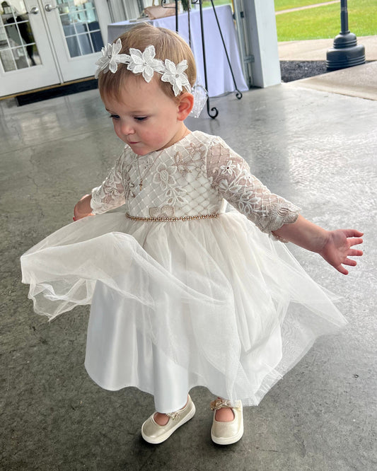 Child in a white lace dress with a floral headband on a concrete floor.
