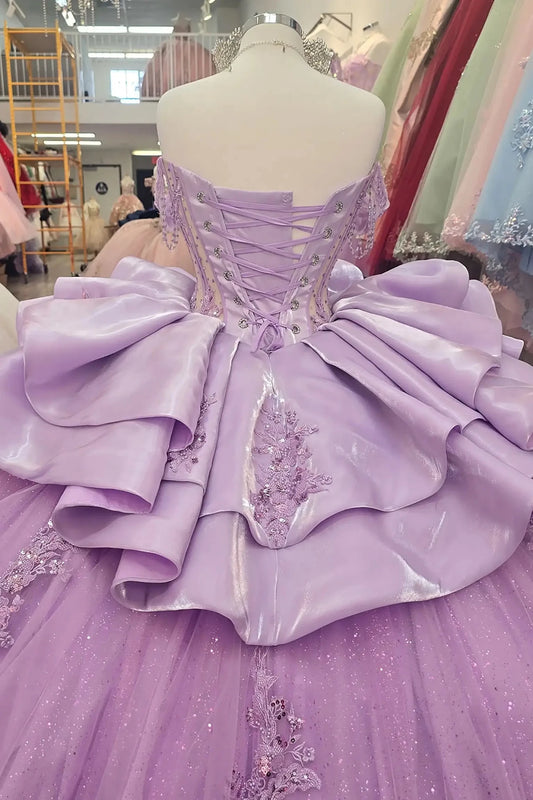 Elegant liliac quinceañera ball gowns on mannequins, featuring beaded sweetheart corset bodices with sheer panels, off-the-shoulder draped sleeves, and adjustable lace-up backs. The ensemble includes sparkling silver tiaras and matching rhinestone necklaces. The gowns showcase voluminous glitter tulle skirts adorned with intricate floral lace appliqués and tiered, sculptural satin ruffles, displayed in a boutique showroom.