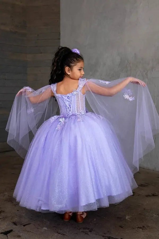 Kid standing back-facing in lilac ball gown with lace-up closure, sheer bodice, dramatic cape, and cascading layered tulle skirt