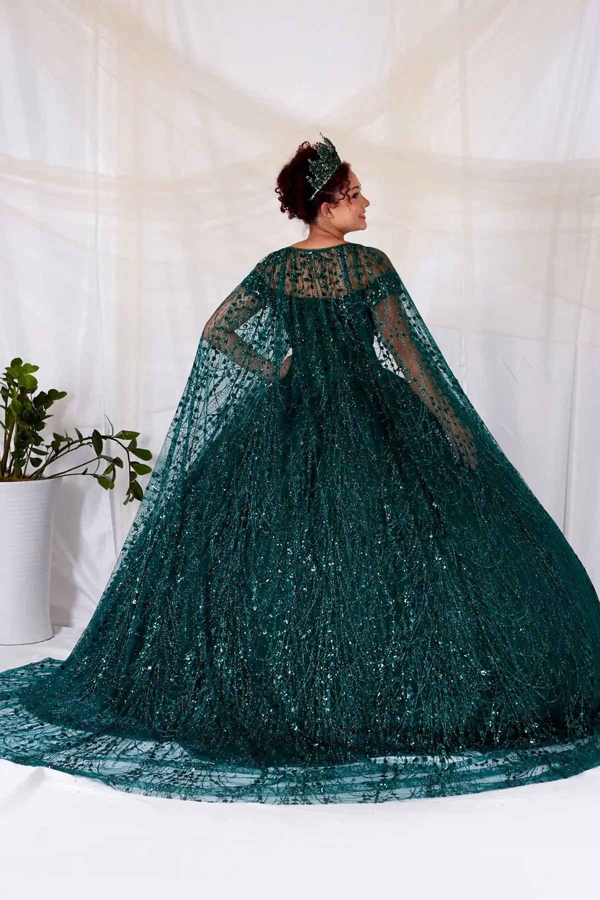 Back-Facing of a woman wearing Emerald formal gown featuring open back design sweeping skirt and attached cape for dramatic effect