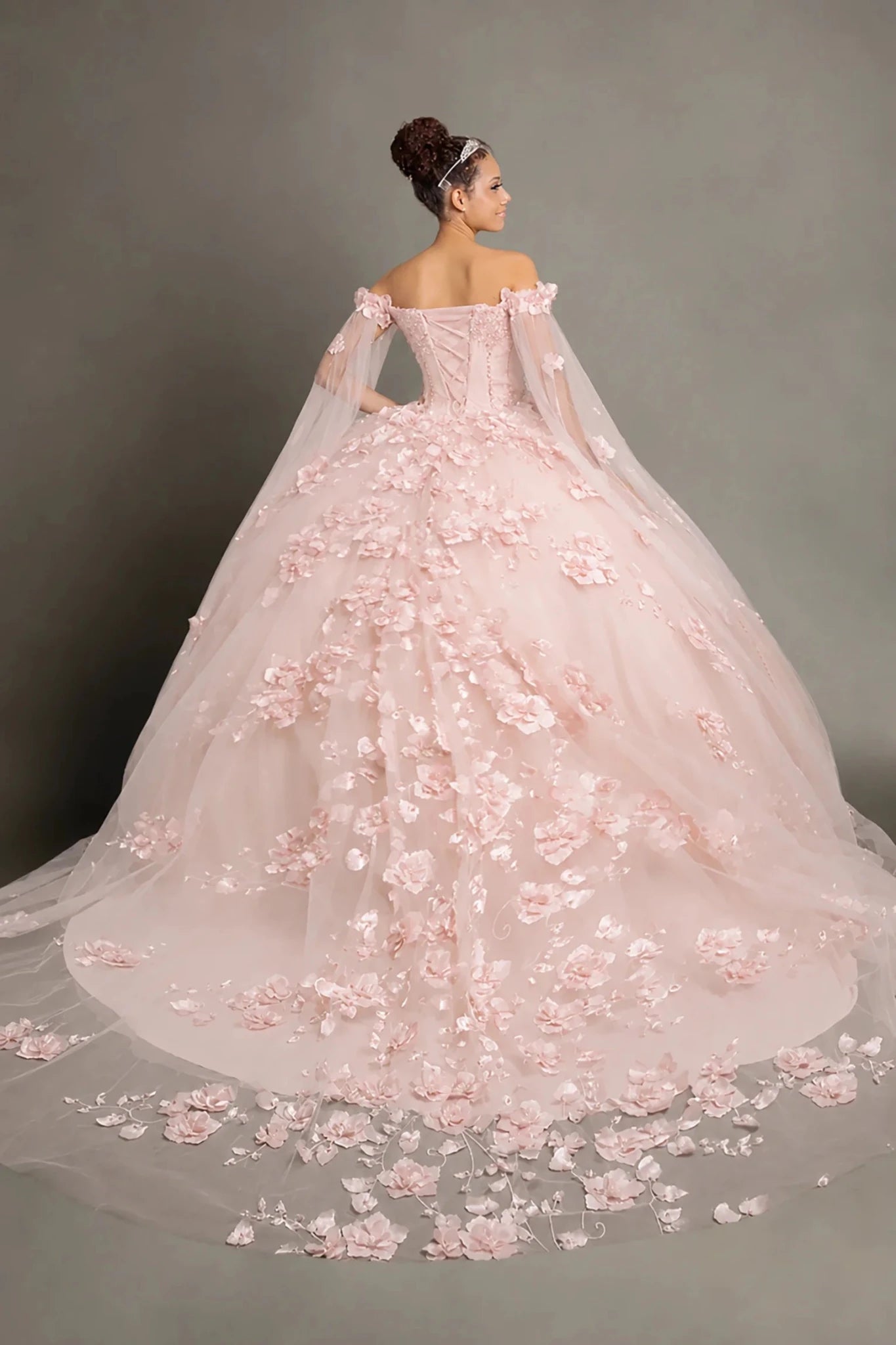 A pale pink quinceanera ball gown with a long sheer cape and voluminous tulle skirt covered in 3D floral appliqué