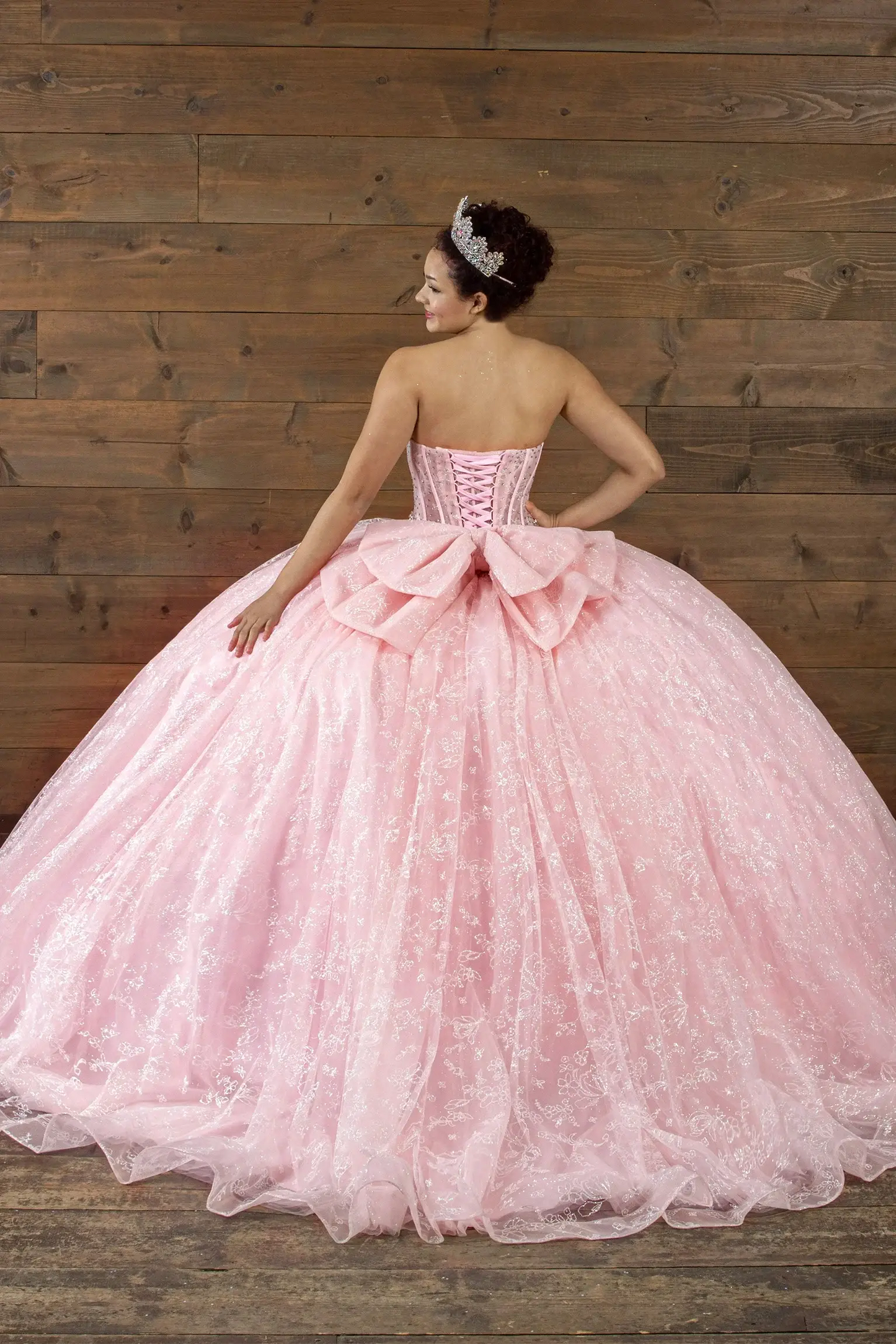 Pink quinceañera ball gown with sheer corset bodice, crystal beading, glitter floral tulle and bow detail