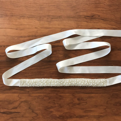 Textured details appear across ivory and white pearl satin flower girl sash belt, close-up, full view, embellishment detail.