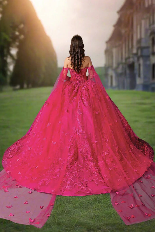 Woman standing back-facing in fuchsia quinceañera ball gown with dramatic off-shoulder design, lace-up beaded bodice, floral embroidery, full tulle skirt, and trailing cape details