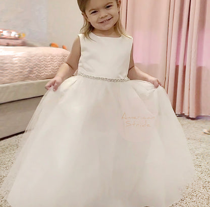 Young girl in a white dress with 'American Stride' branding, standing on a carpeted floor.