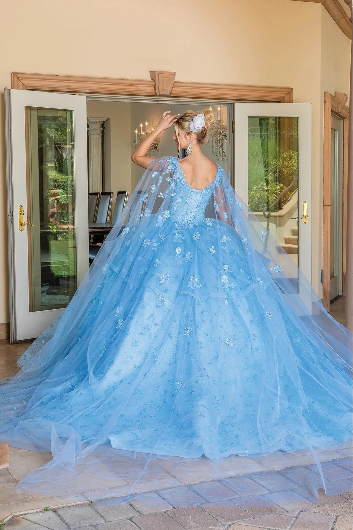 Back view of a Bahama Blue quinceañera gown showing lace-up back, sheer cape, and floral appliqués