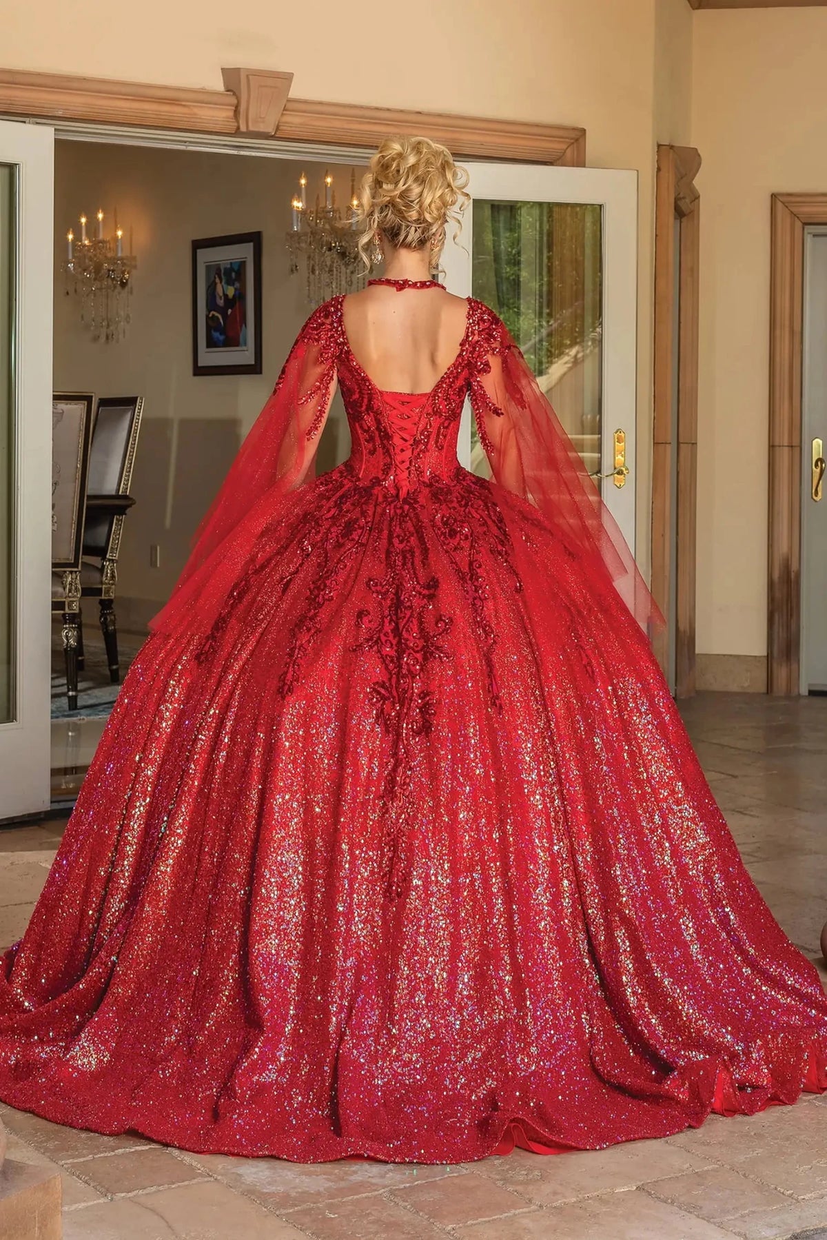 Back view of a Red quinceañera gown showing lace-up back, dramatic sheer cape, and sequined lace
