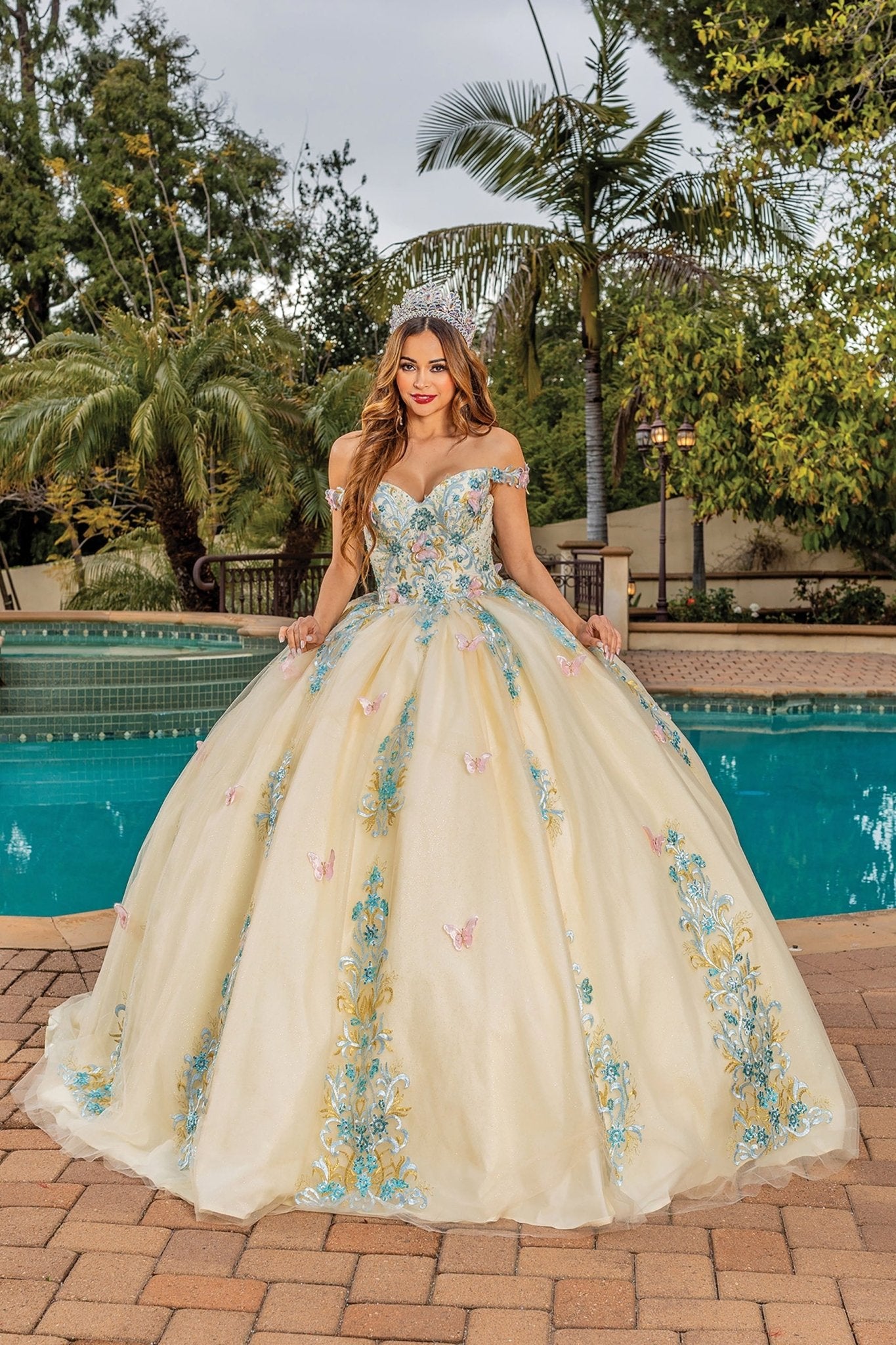 Quinceanera | Off-the-Shoulder Crystal Embellished Lace Appliqués, Beaded Butterflies, and Tulle Skirt Quinceanera dress - front view of woman wearing dress standing in front of pool