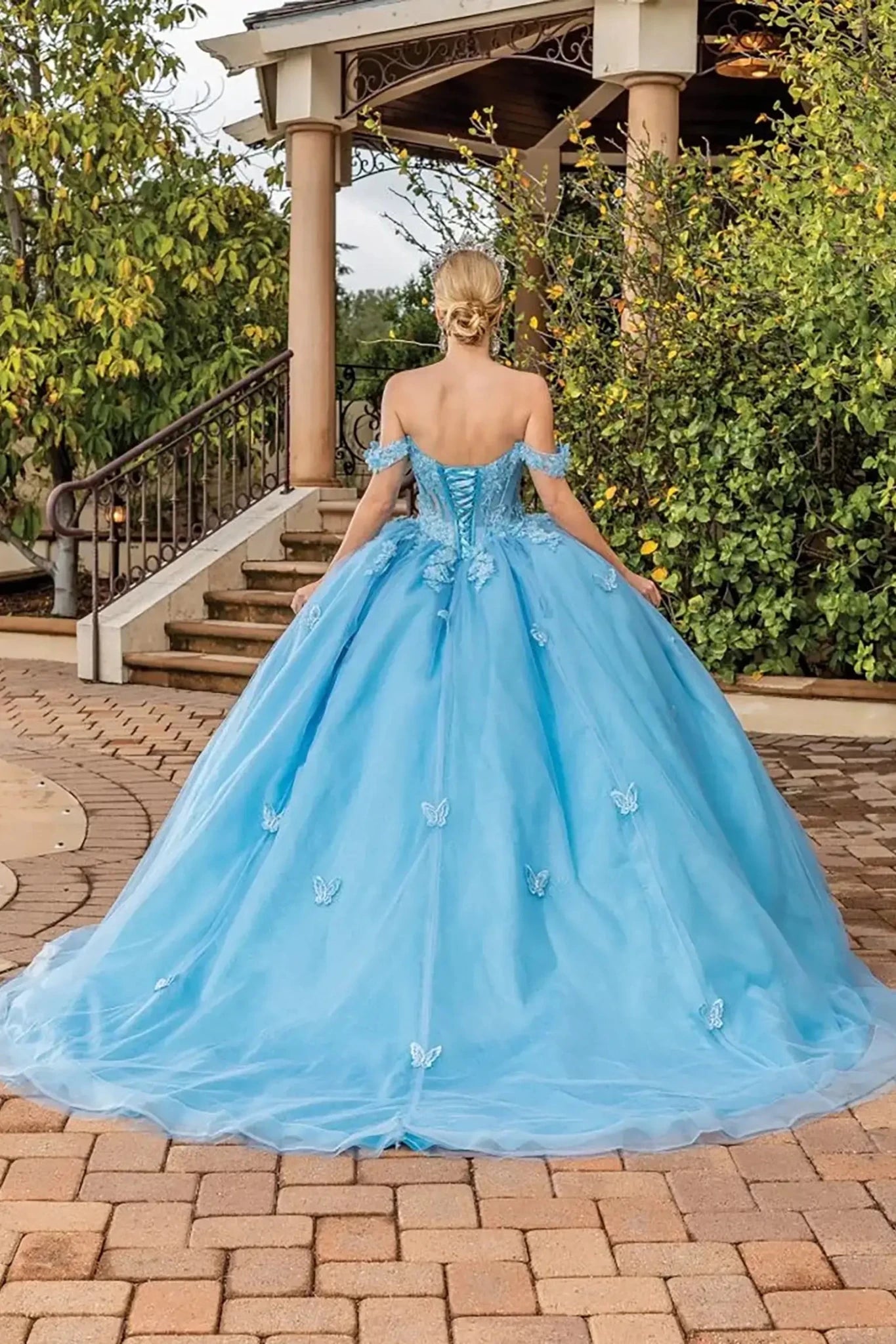 Back view of a Bahama blue off-shoulder quinceañera ball gown featuring a lace-up corset back, butterfly appliqués, flowing glitter tulle skirt, and full train.