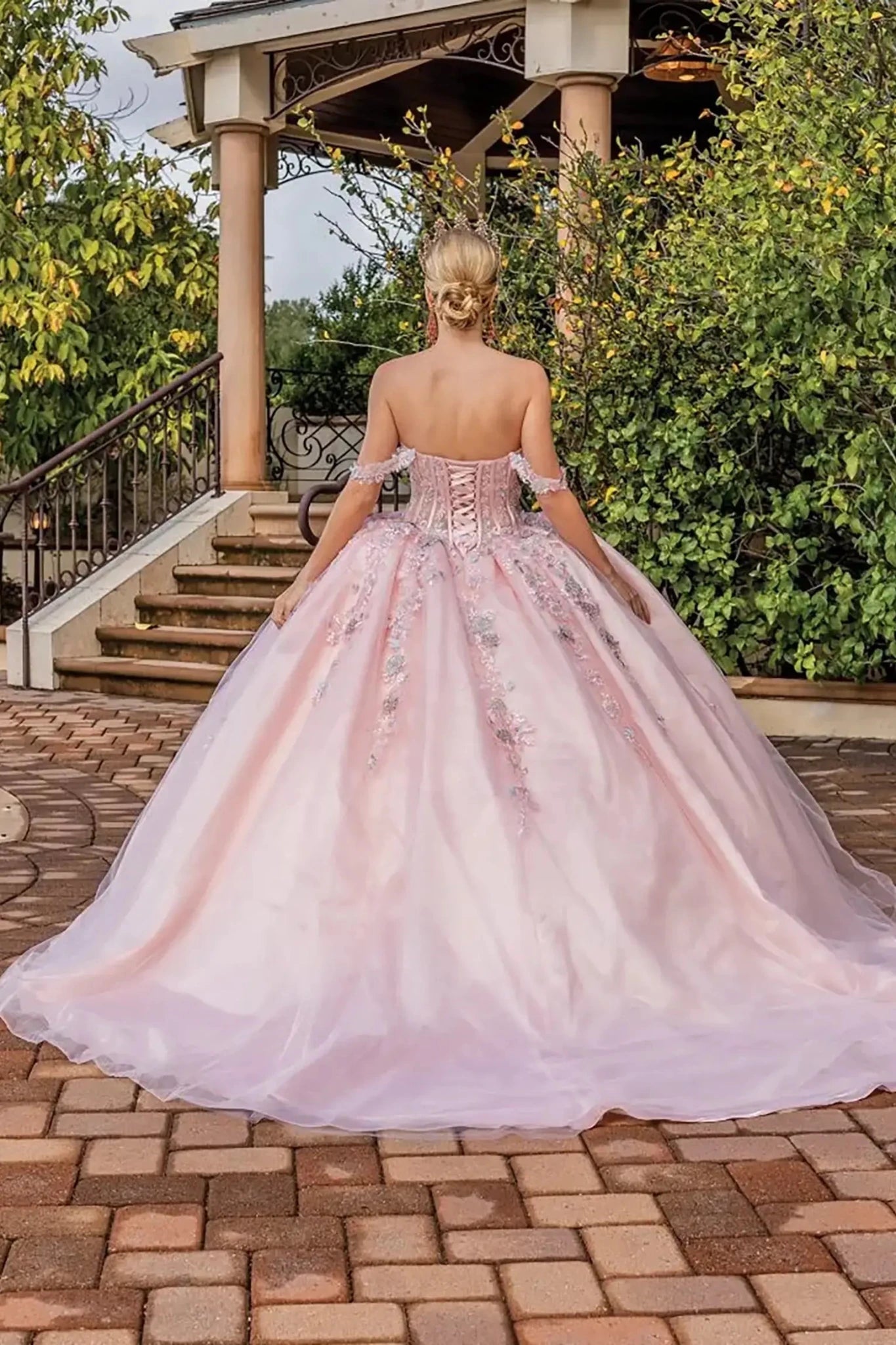 Back view of a blush pink off-shoulder quinceañera ball gown featuring a lace-up corset back, floral appliqués, flowing tulle skirt, and soft train.