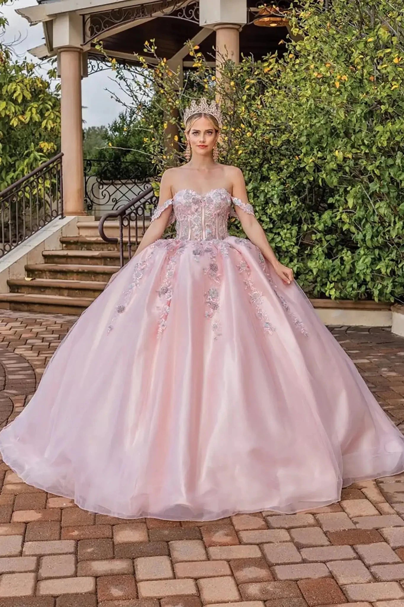 Front view of a blush pink off-shoulder quinceañera ball gown with sheer corset bodice, floral appliqués, sparkling embellishments, and a full tulle skirt.