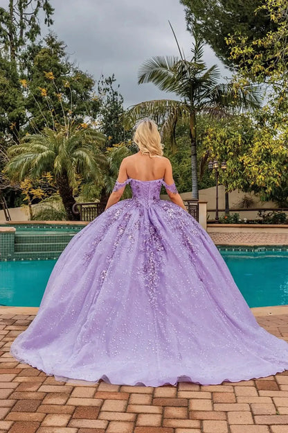 Back view of a lilac off-shoulder quinceañera ball gown featuring a lace-up corset back, shimmering glitter tulle skirt, flowing train, and elegant outdoor setting.