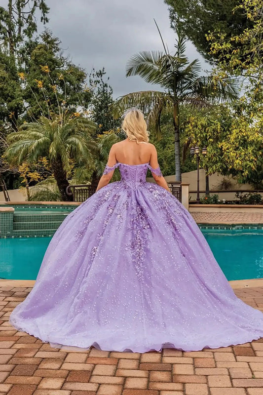 Back view of a lilac off-shoulder quinceañera ball gown featuring a lace-up corset back, shimmering glitter tulle skirt, flowing train, and elegant outdoor setting.