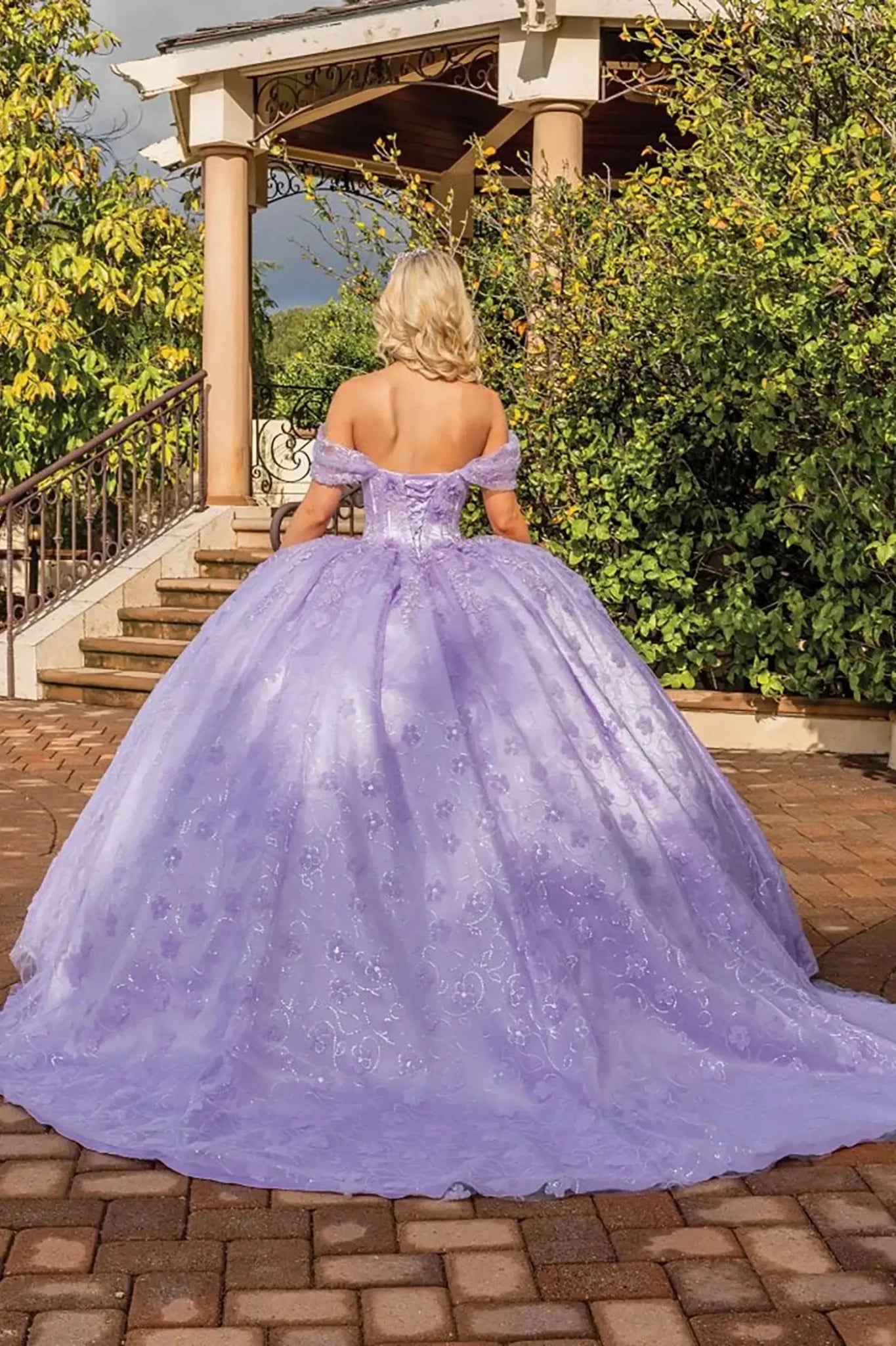 Back view of a lilac off-shoulder quinceañera ball gown featuring a fitted corset back, embroidered glitter tulle skirt, flowing train, and elegant outdoor venue backdrop.