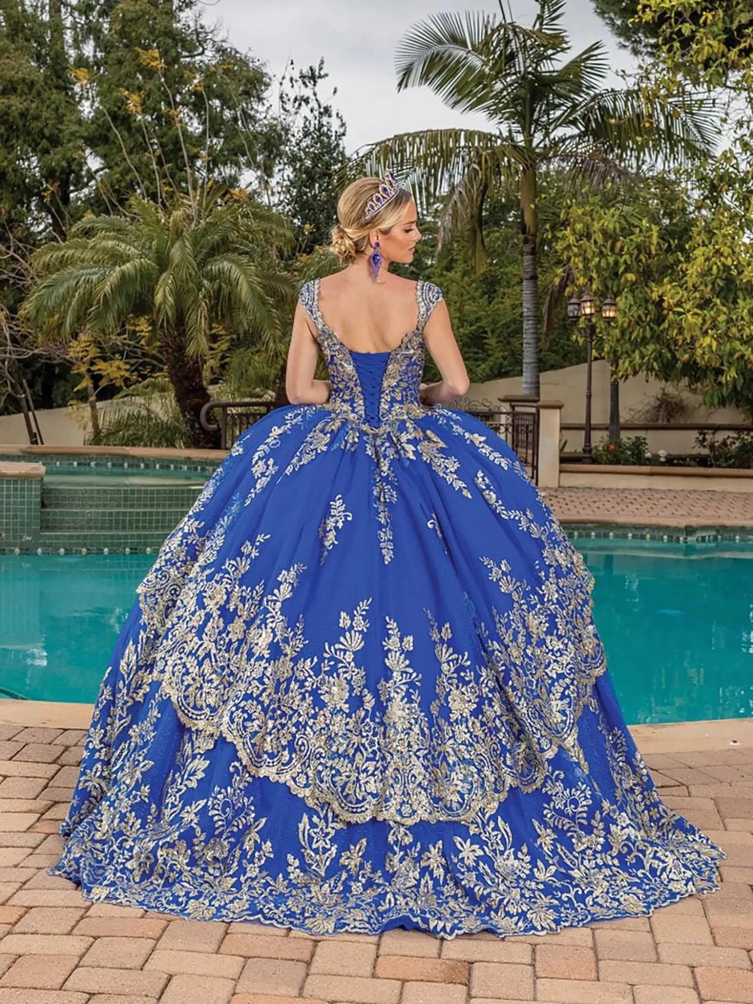 Back view of royal blue quinceañera ball gown showcasing deep V-back corset, ornate gold embroidery, voluminous ball gown skirt, and elegant silhouette photographed outdoors by a pool