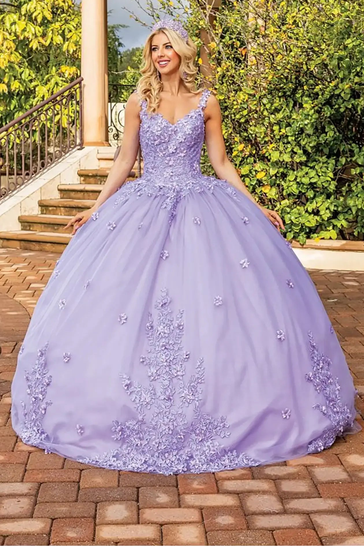 Front view of a lilac quinceañera ball gown with sleeveless embellished corset bodice, delicate floral appliqués, layered tulle skirt, and a full princess silhouette in an outdoor garden setting.