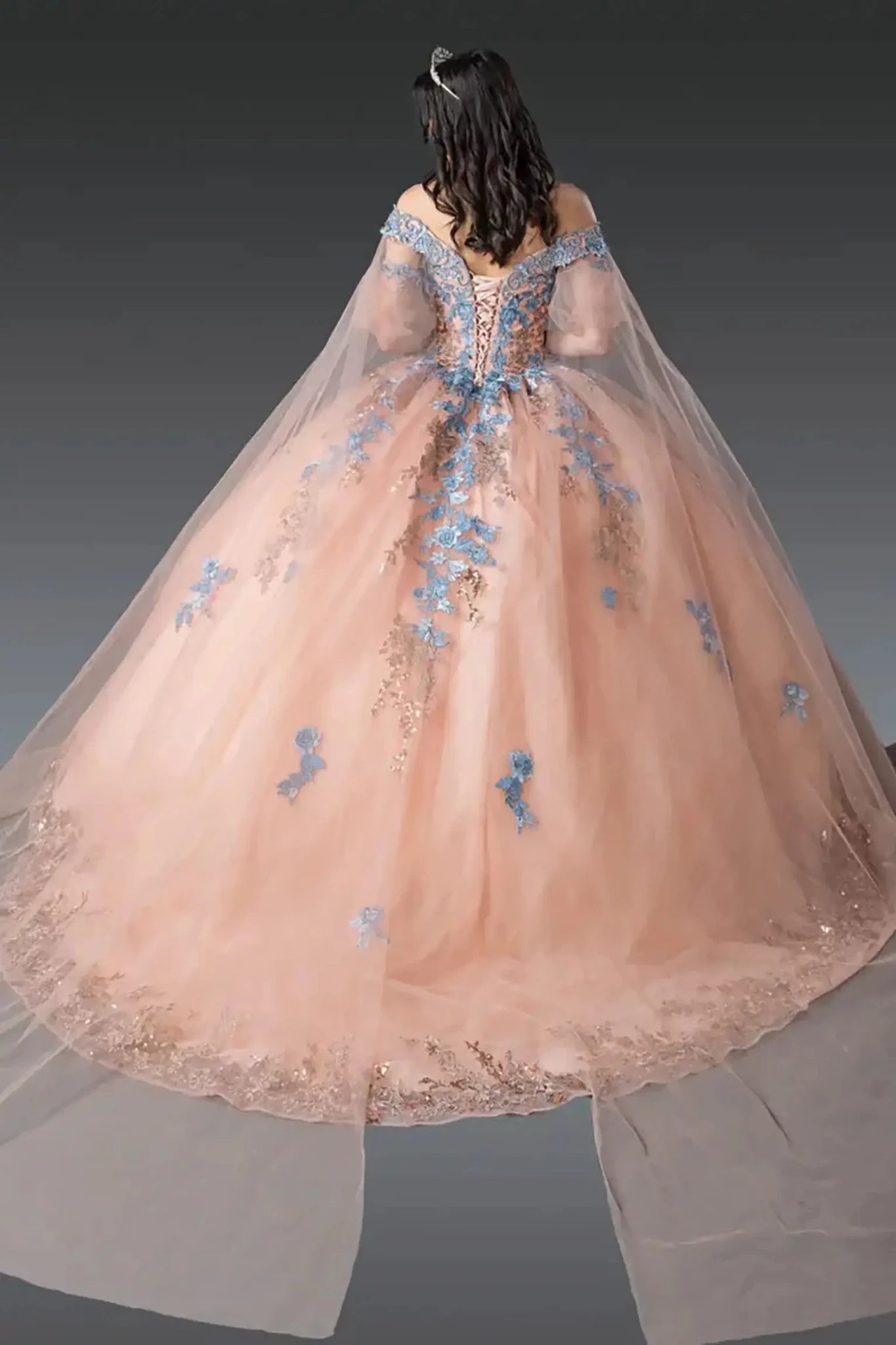 Back view of rose gold quinceañera ball gown showcasing lace-up corset back, sheer cape-style tulle overlay, blue floral embroidery, and full ball gown silhouette photographed in studio lighting