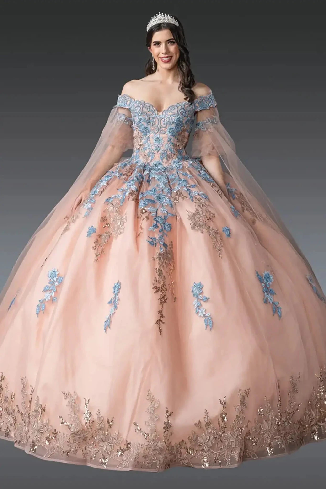 Front view of rose gold quinceañera ball gown featuring off-shoulder embroidered corset bodice, sheer tulle sleeves, blue floral appliqués, layered tulle skirt, and tiara styling photographed in studio