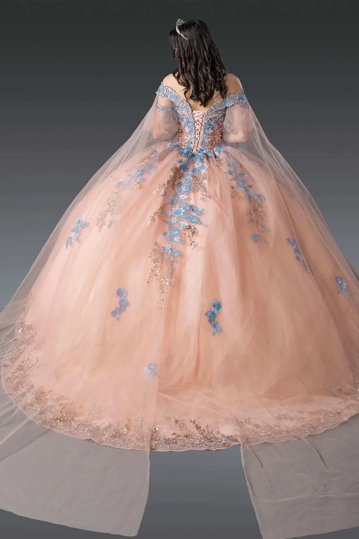 A voluminous rose gold tulle quince ball gown shown from the back on a model, featuring light blue floral embroidery, a lace-up corset-style back, and a sheer tulle cape train 