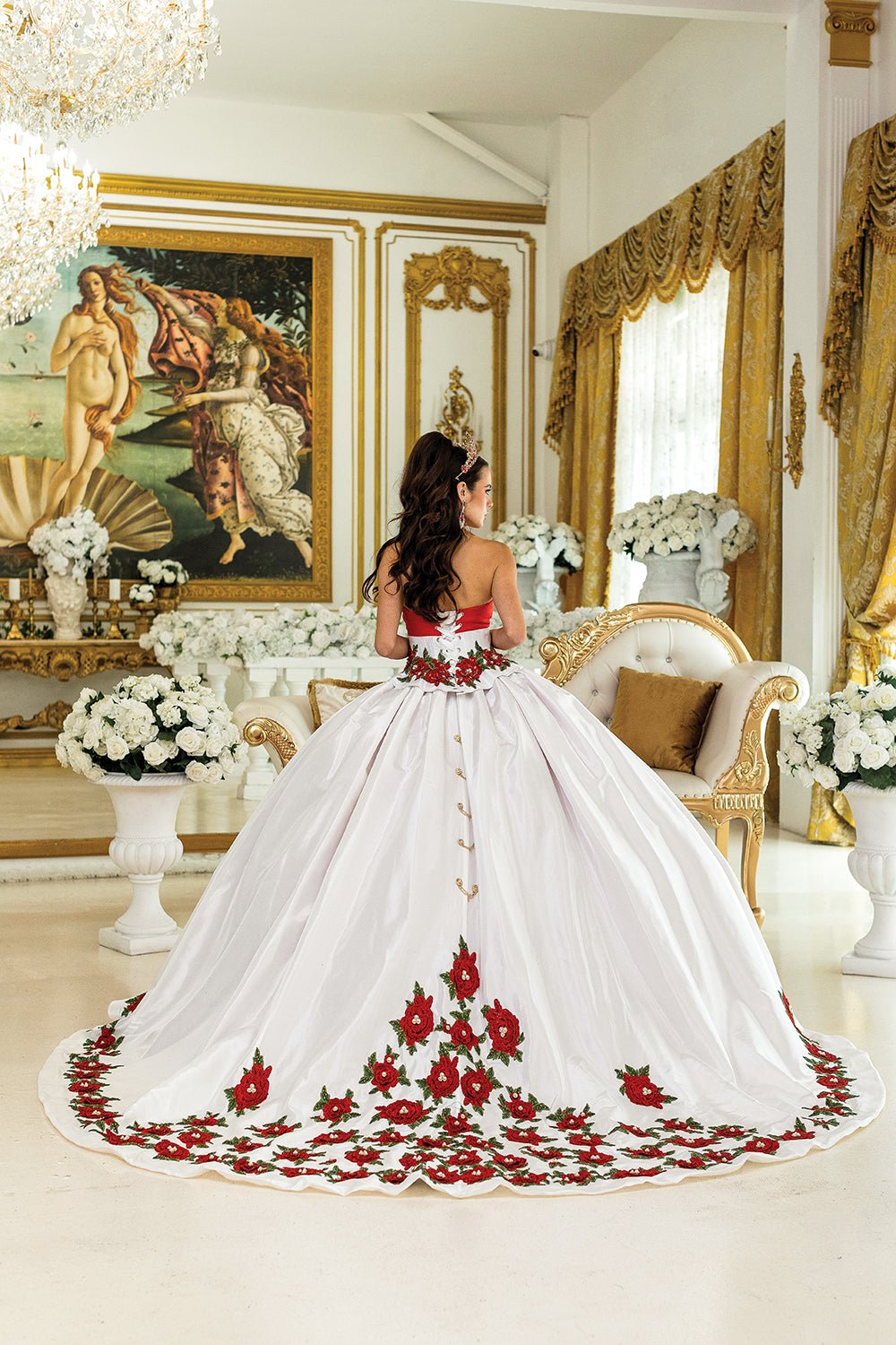 Woman standing back-facing in white and red strapless quinceañera ball gown with lace-up embroidered floral bodice, layered ruffled skirt, and butterfly accents on train