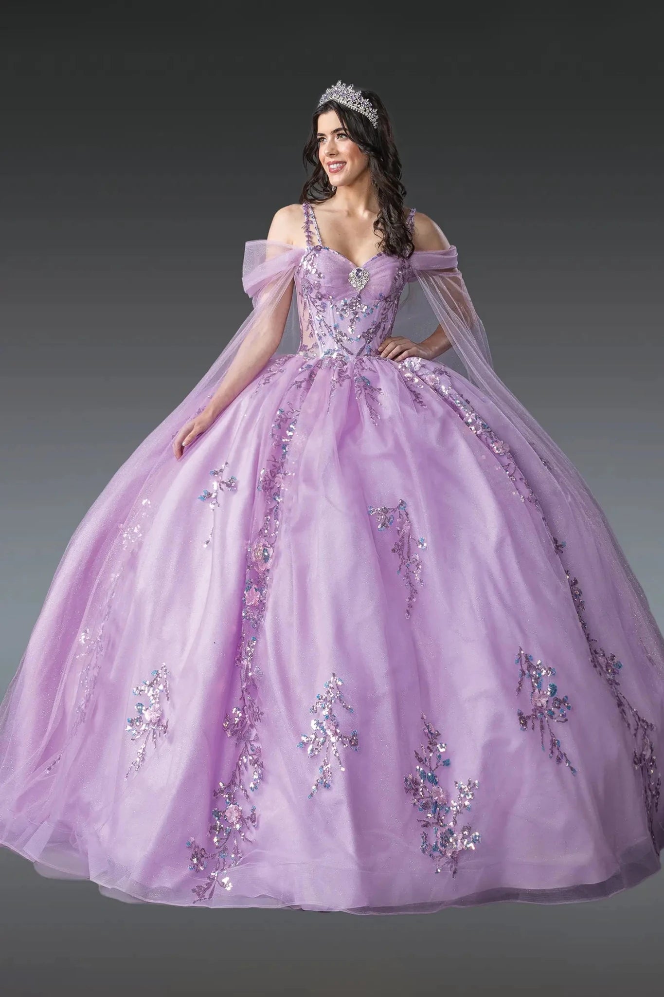 Woman standing front-facing in lilac quinceañera ball gown with sheer puffed sleeves, fitted bodice, embellished floral appliqués, and voluminous layered tulle skirt