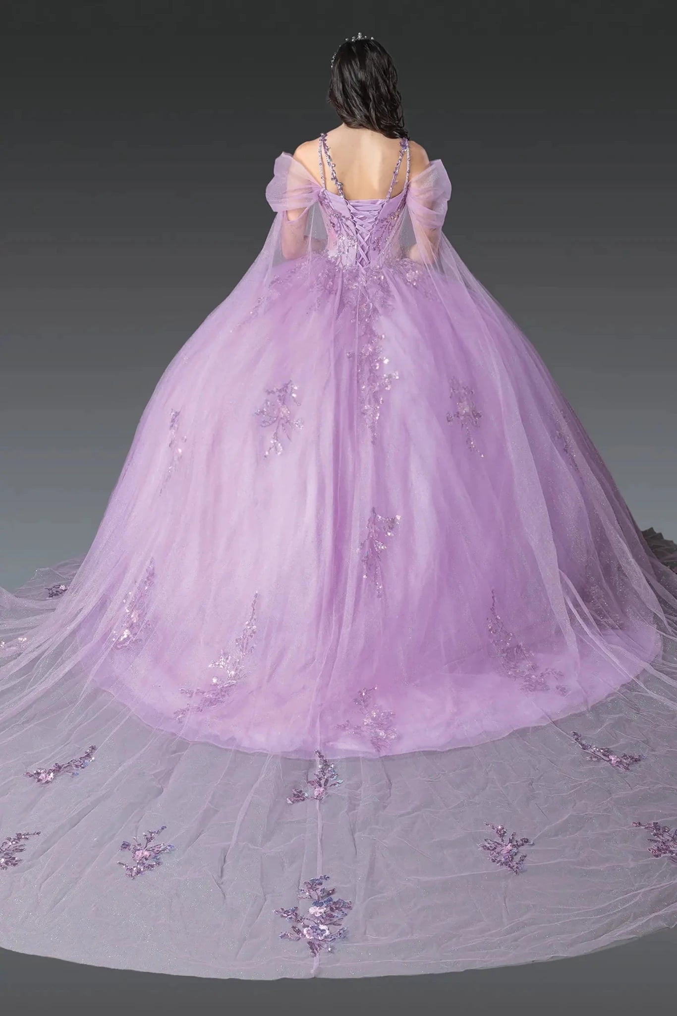 Woman standing back-facing in lilac quinceañera ball gown with lace-up bodice, sheer puffed sleeves, floral appliqués, and flowing layered tulle skirt with train