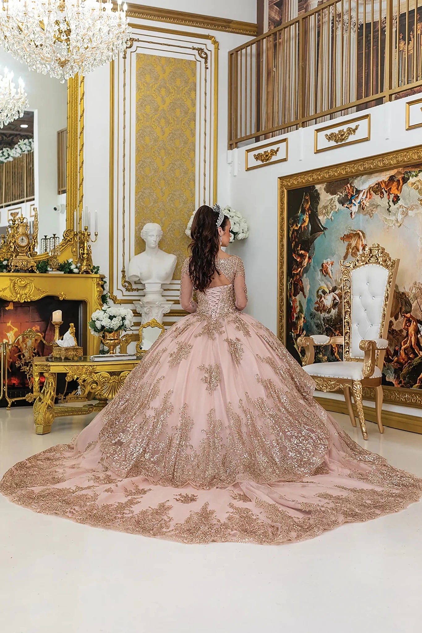 Woman standing back-facing in elegant rose gold quinceañera ball gown with lace overlay, lace-up bodice, sheer long sleeves, and flowing sparkling skirt with train