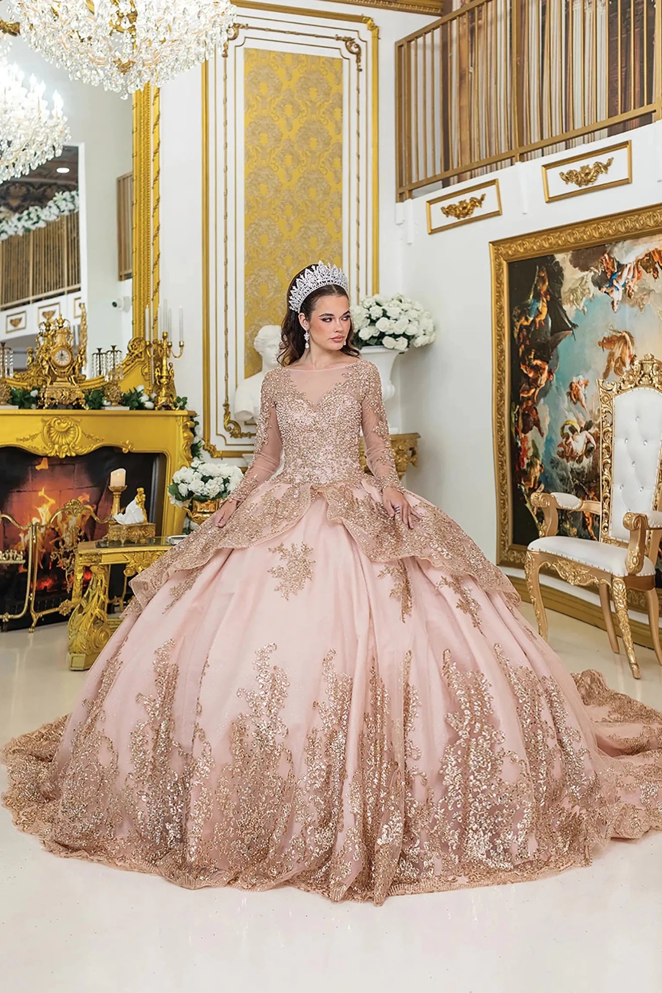 Woman standing front-facing in elegant rose gold quinceañera ball gown with lace overlay, sheer long sleeves, fitted bodice, and layered sparkling skirt