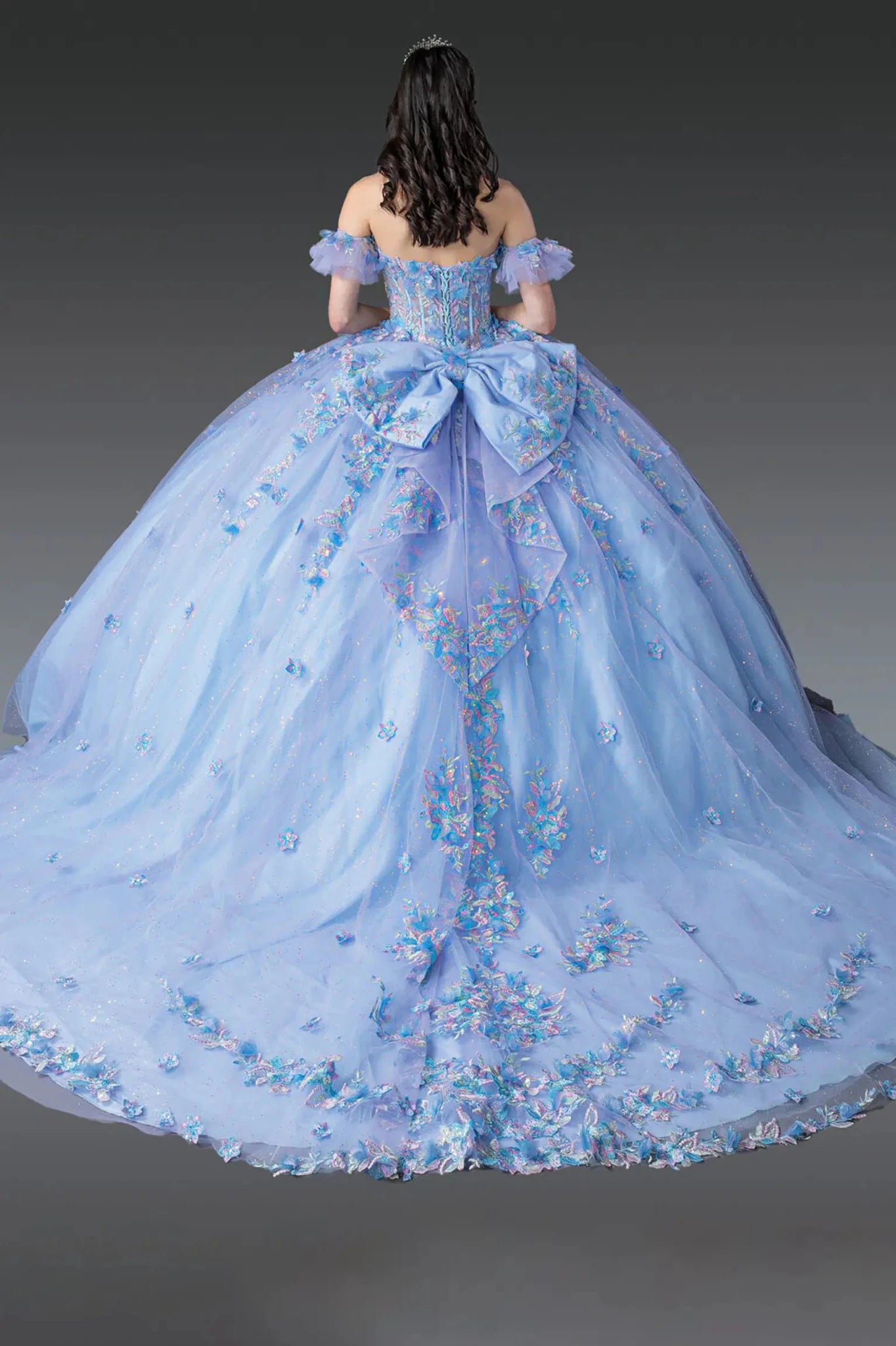 Woman standing back-facing in Bahama blue-lilac quinceañera ball gown with lace-up beaded bodice, off-shoulder sleeves, floral appliqués, and flowing embellished tulle skirt with train